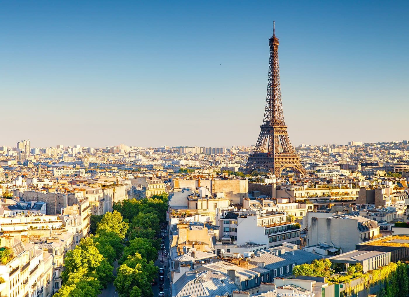 Eiffel Tower rising above Paris cityscape with tree-lined streets and buildings under a clear blue sky.